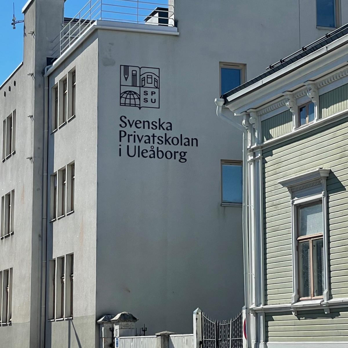 Det gråa Weckmanska huset som tillhör Svenska Privatskolan i Uleåborg syns till vänster. Stenhuset i funkisstil har skolans namn och logo på väggen på den tredje och översta våningens nivå. Till höger syns en del av ett ljusgrönt envåningshus i trä med en hög stengrund, Snellmanska huset från 1800-talet. Mellan husen syns en port mot skolans gård. Vyn är söderut längs Kyrkogatan i Uleåborg. Fotot är taget 2024 av Tea Stolt de Glanville, lärare vid skolan.