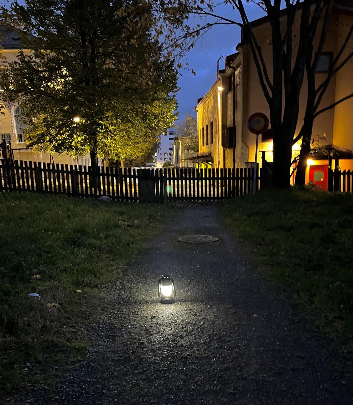 En upplyst lykta på en mörk gångstig mellan träd och byggnader en höstkväll.