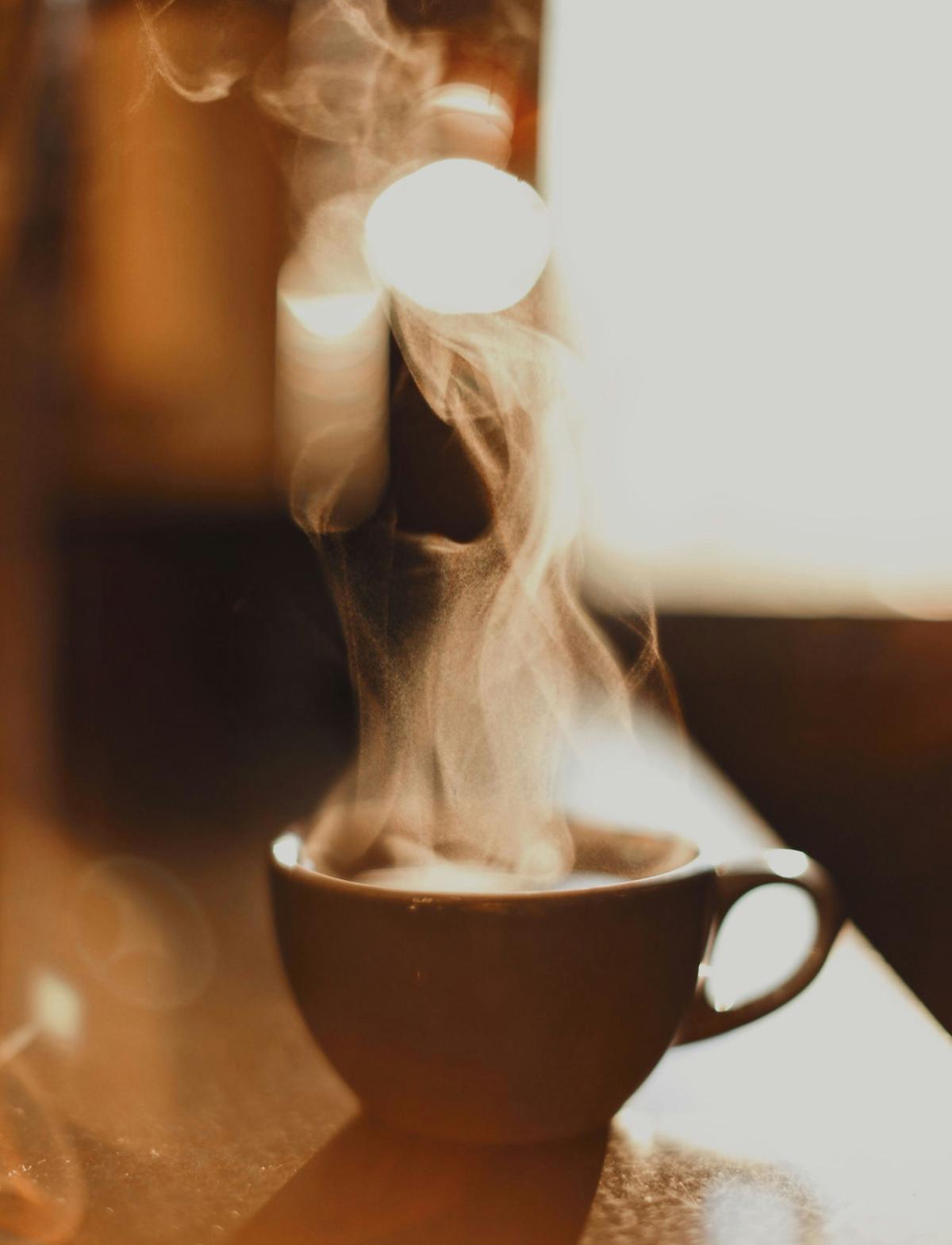 En mörk kaffekopp står på ett bord i varm belysning. Tunna stråk av ånga stiger upp från den varma drycken och formar mjuka, slingrande mönster mot den oskarpa bakgrunden. Bilden domineras av varma bruna och gyllene toner som skapar en inbjudande och stillsam atmosfär.