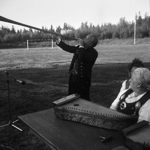 Person spelar ett långt traditionellt blåsinstrument utomhus. I förgrunden syns ett stränginstrument på ett bord och personer i folkdräkt.