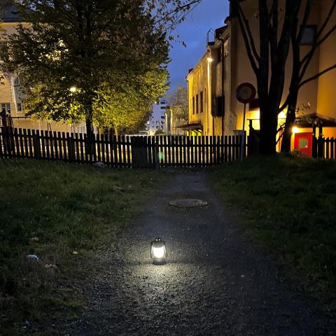 En upplyst lykta på en mörk gångstig mellan träd och byggnader en höstkväll.