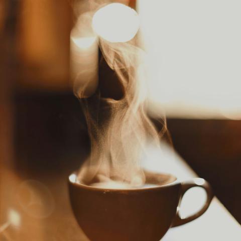En mörk kaffekopp står på ett bord i varm belysning. Tunna stråk av ånga stiger upp från den varma drycken och formar mjuka, slingrande mönster mot den oskarpa bakgrunden. Bilden domineras av varma bruna och gyllene toner som skapar en inbjudande och stillsam atmosfär.